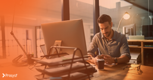 Colaborador sentado em frente à uma mesa de trabalho, conferindo suas notícias no celular enquanto acompanha seu computador.